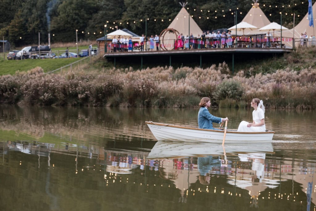 A couple in wedding attire is rowing a small boat on a calm lake. In the background, there are guests at an outdoor wedding reception under string lights, with several tents on a grassy hill.