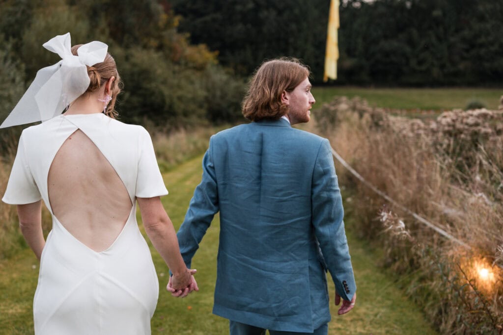 A couple, seen from behind, walks hand in hand on a grassy path. The woman wears a white dress with an open back and a bow in her hair, while the man wears a blue suit. They are surrounded by greenery and softly lit tall grass.