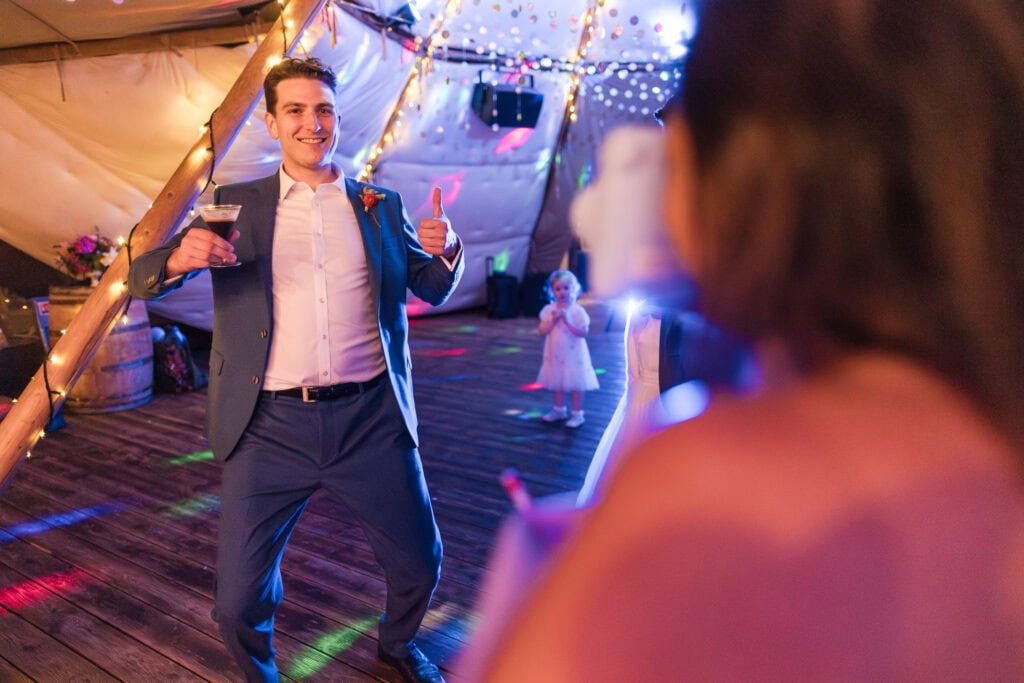 A man in a suit enthusiastically gives a thumbs-up while holding a drink on a dance floor, illuminated by colorful lights. A child in the background observes, and a woman's back is visible in the foreground.