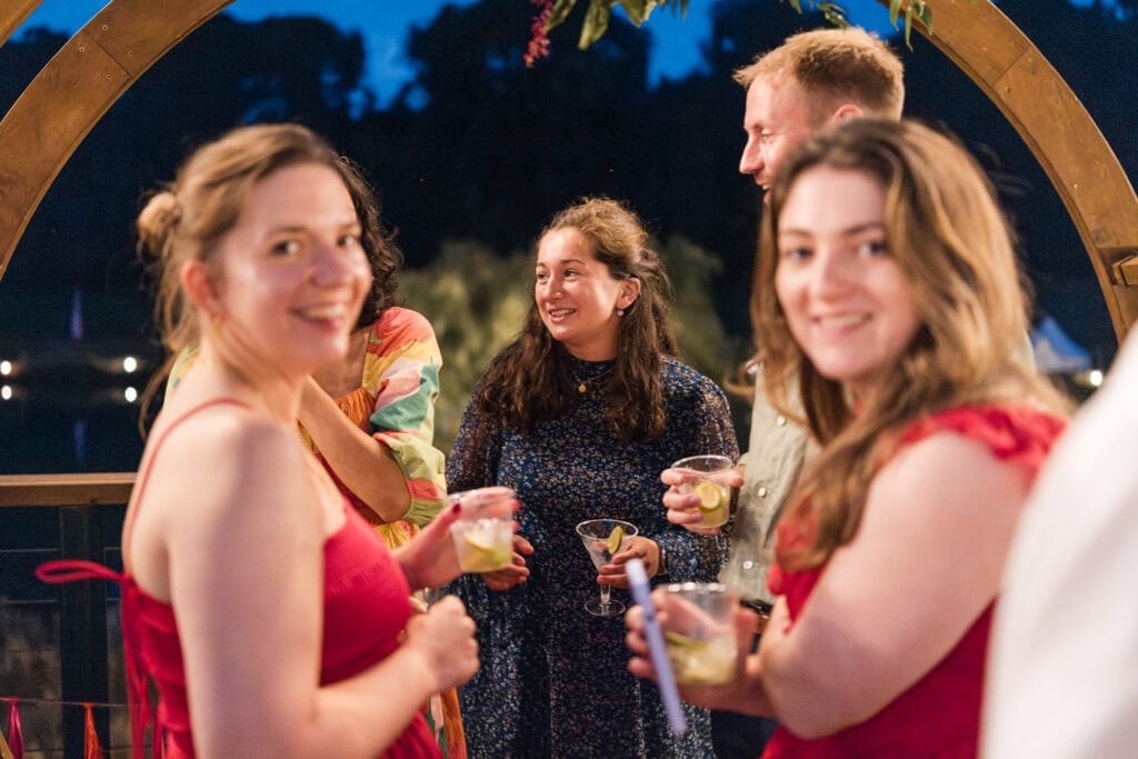 A group of people socializing and holding drinks at an outdoor event in the evening. They are smiling and engaging in conversation, with trees and a dark blue sky in the background.