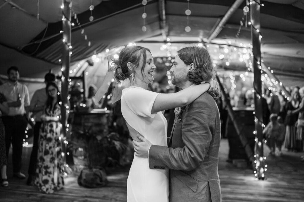 A bride and groom share their first dance in a warmly lit venue adorned with string lights. They embrace happily, surrounded by blurred guests watching in the background. The atmosphere is joyful and intimate.