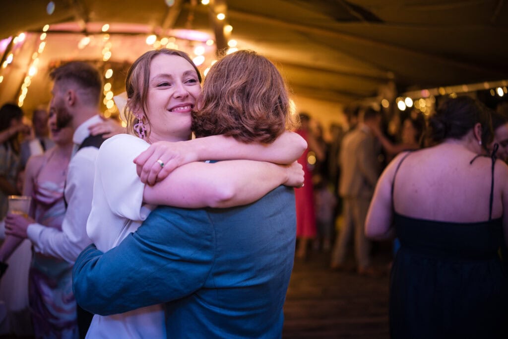 A person in a white dress hugs someone in a blue jacket on a dance floor, surrounded by people and colorful lights. The atmosphere is lively and joyful.