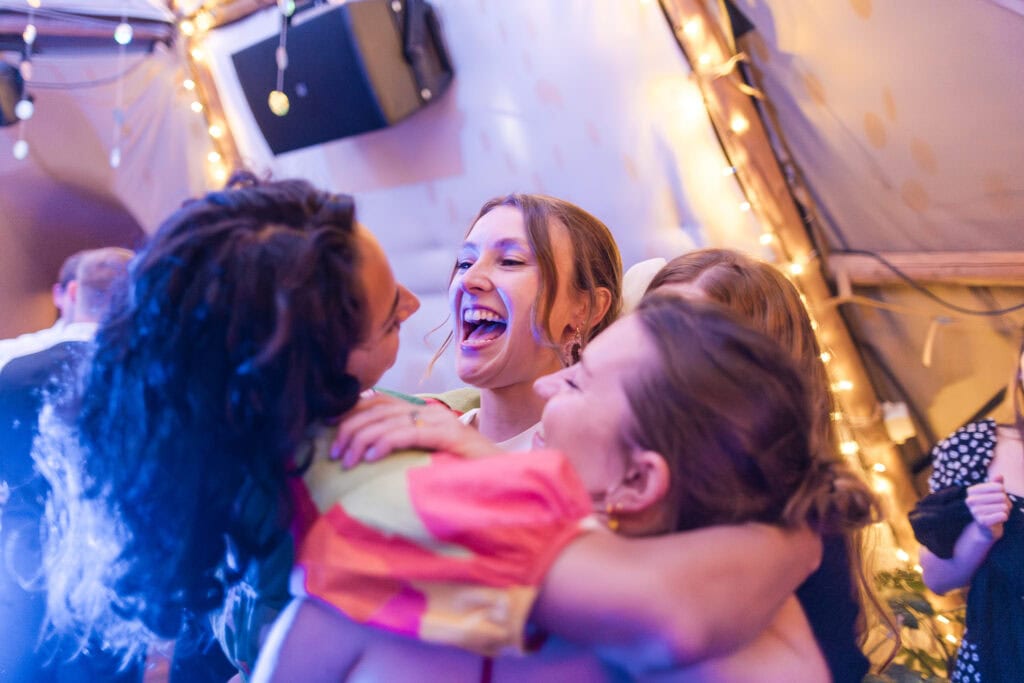 A group of four women embracing and laughing joyfully under warm festive lighting. The setting has decorative string lights, creating a lively and celebratory atmosphere.