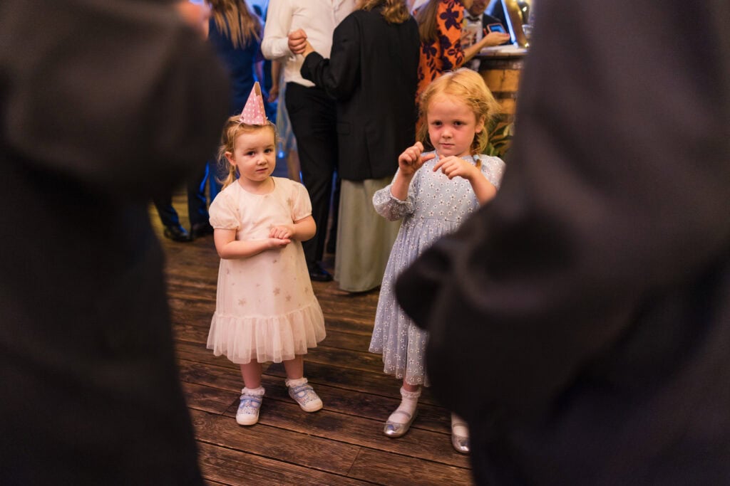 Two young girls stand indoors. One wears a light pink dress and party hat, while the other wears a light blue dress. They are surrounded by adults seen in partial view. The setting appears to be a lively social gathering.