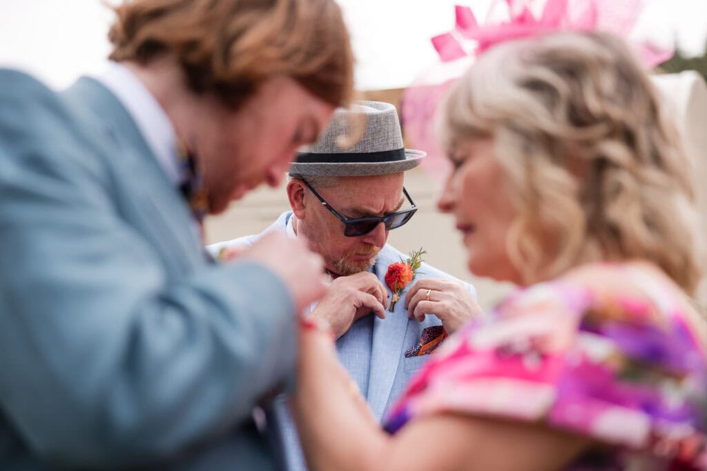 A man wearing sunglasses and a hat is adjusting a flower on his light blue jacket at an event. Two other people, a woman in a floral dress and a man in a blue suit, are standing nearby. They are focused on their attire.