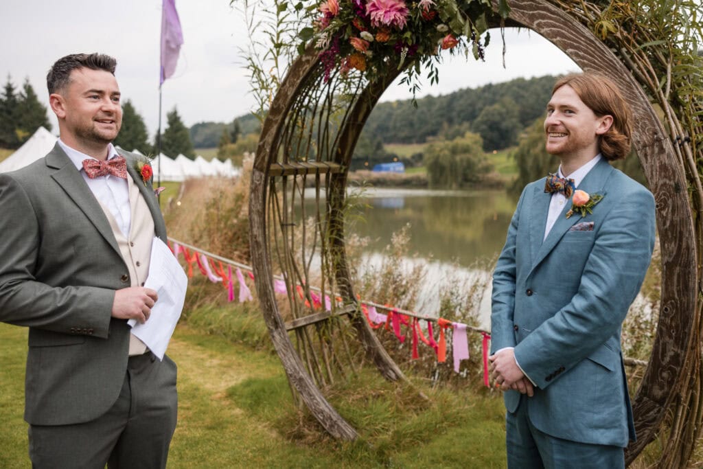 Two people standing outdoors near a floral arch by a lake, dressed in suits with bow ties. One is holding papers and speaking, while the other is smiling. Tents and trees are visible in the background.