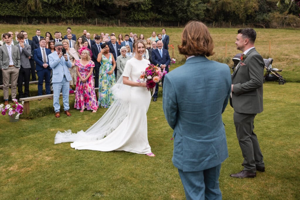 A bride in a white gown and veil stands on a grassy lawn, holding a bouquet, facing the groom in a dark suit. A man in a blue suit stands nearby. Guests seated and standing in colorful outfits watch from the background.