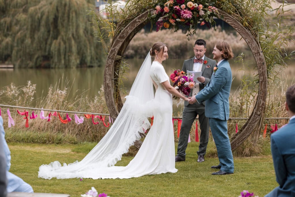 A bride and groom stand facing each other in front of a floral arch by a lake. The bride holds a vibrant bouquet, and a person stands between them, officiating. Guests are seated, watching the outdoor ceremony.