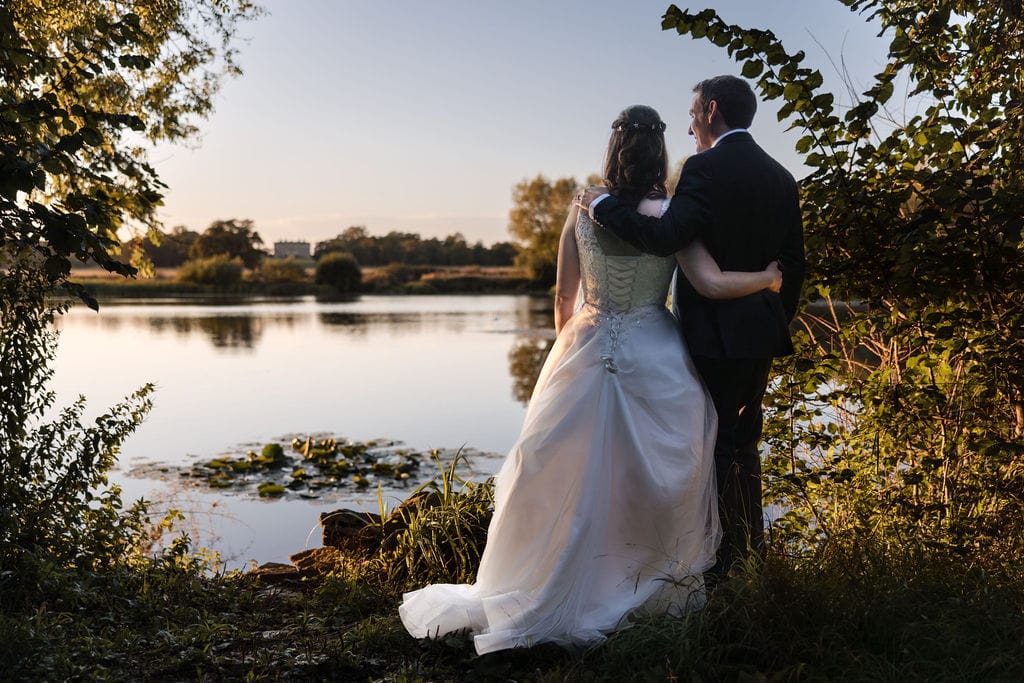 Jenn-Martyn-303 A couple in wedding attire embraces as they stand by a tranquil lake, surrounded by greenery. The setting sun casts a warm glow, creating a serene and romantic atmosphere.