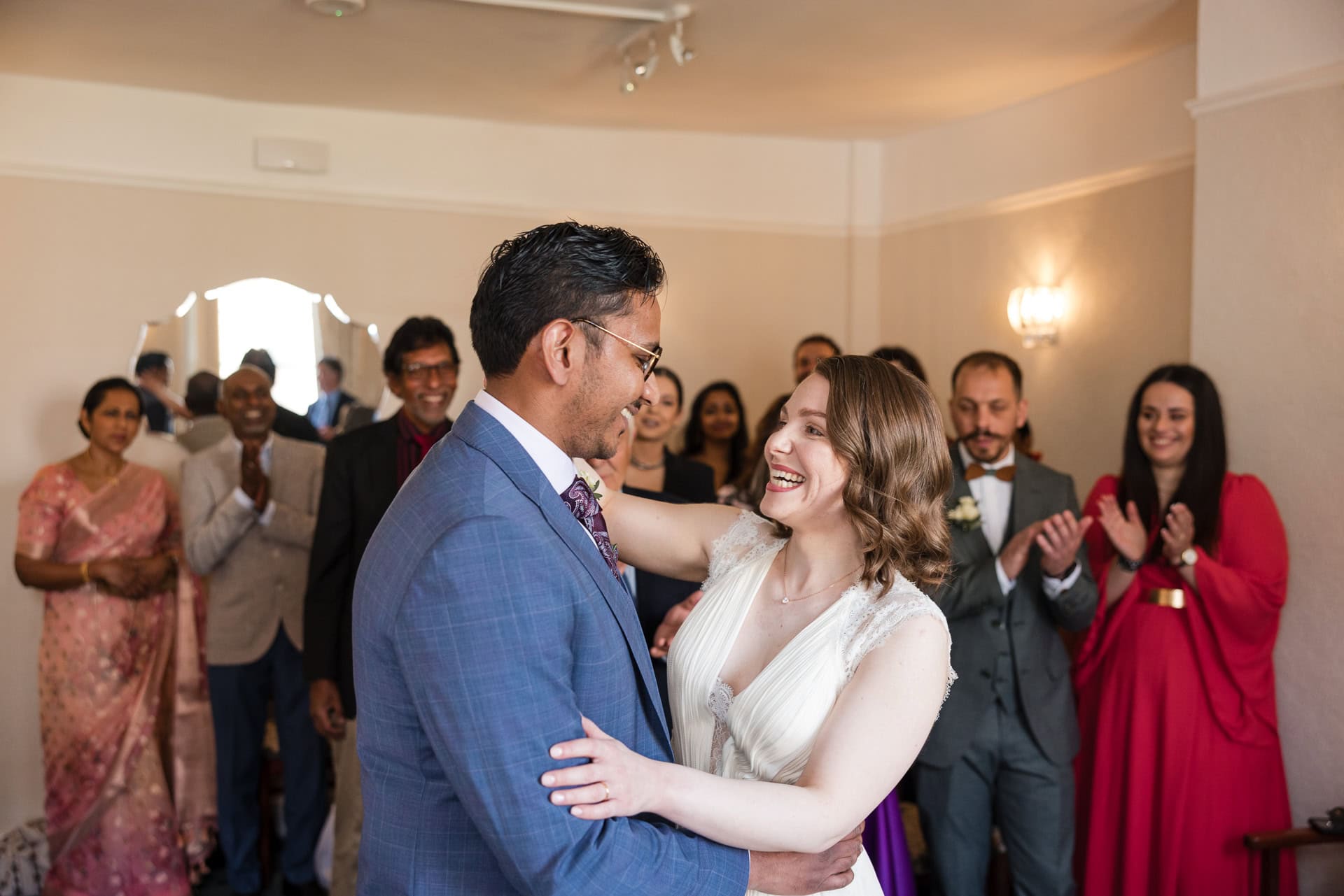 A bride and groom share a joyful first dance in a warmly lit room, surrounded by applauding guests. The bride wears a white dress and the groom wears a blue suit. Friends and family stand around, some in colorful attire, clapping and smiling.