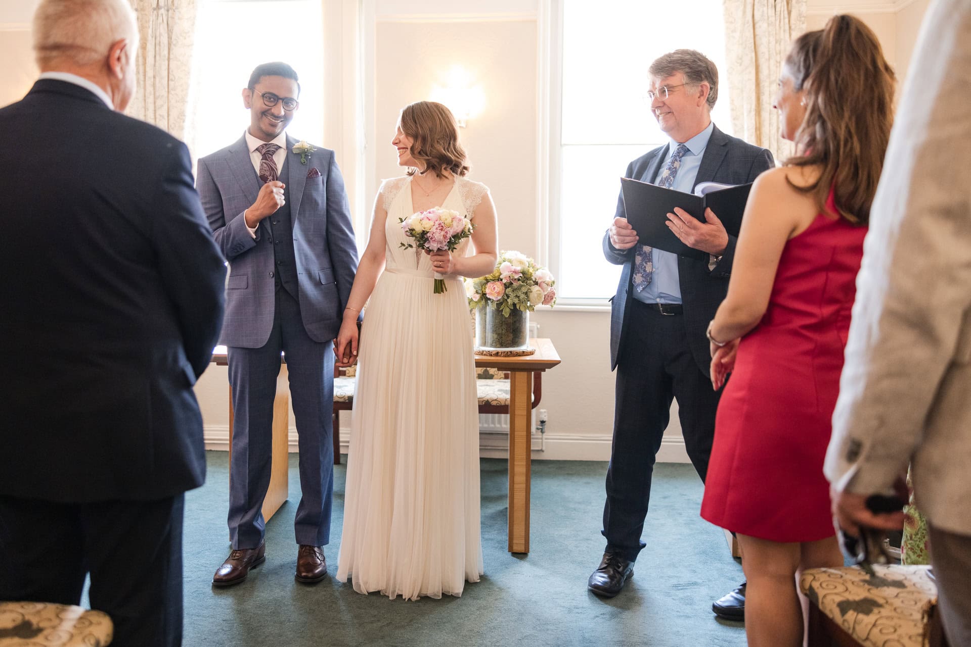 A couple stands at the altar, holding hands during a wedding ceremony. The groom is in a suit, and the bride is in a white dress holding a bouquet. An officiant with a book stands beside them, with guests observing.