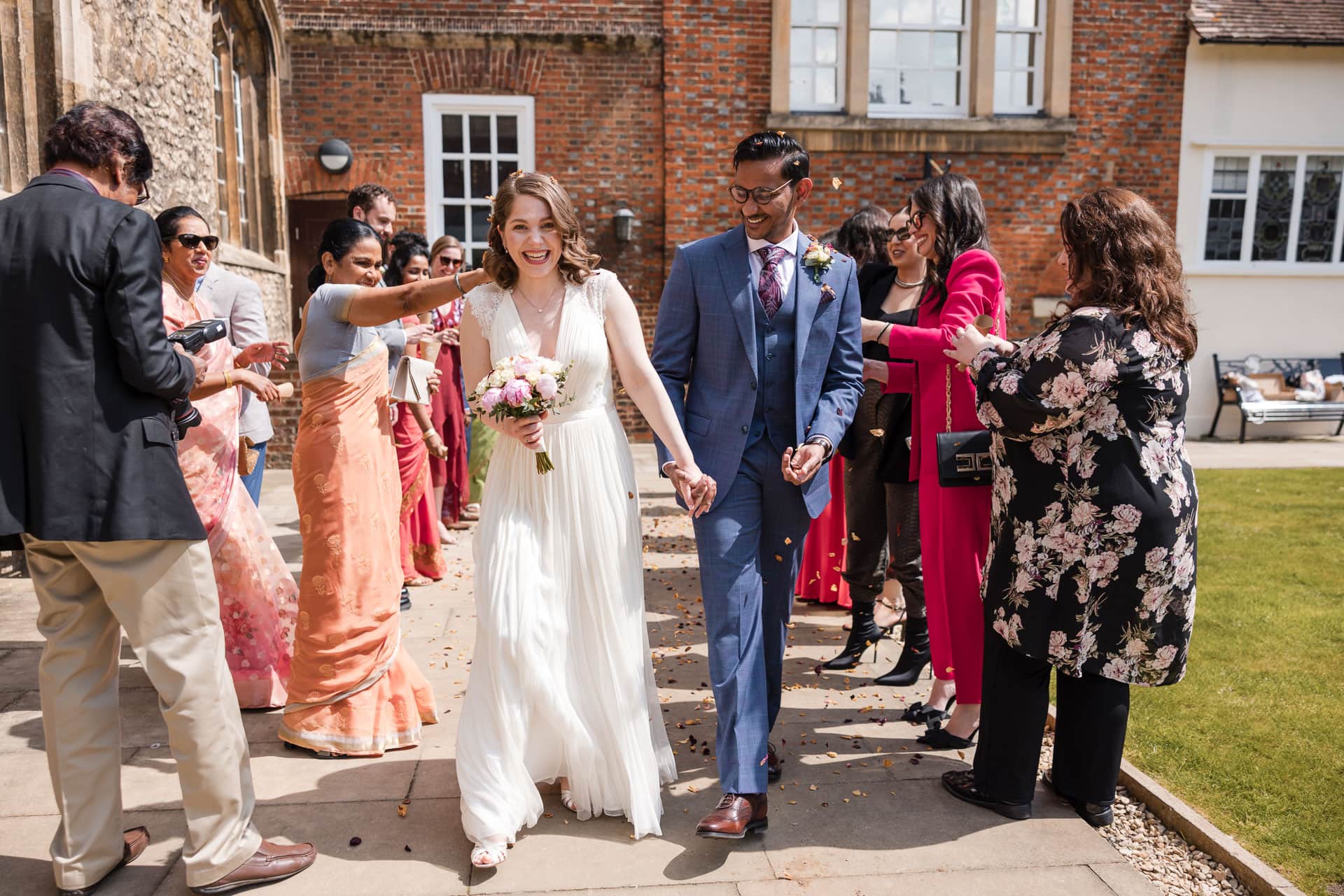 A newlywed couple, the bride in a white dress holding a bouquet, and the groom in a blue suit, walk hand in hand while guests cheer and take photos outside a brick building on a sunny day.
Oxford Wedding Photographer