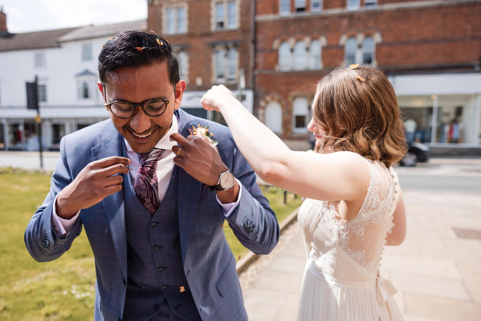 A man in a blue suit and tie adjusts his attire while smiling. A woman in a white dress joyfully helps him. They are outdoors on a sunny day, with brick buildings and a grassy area in the background.