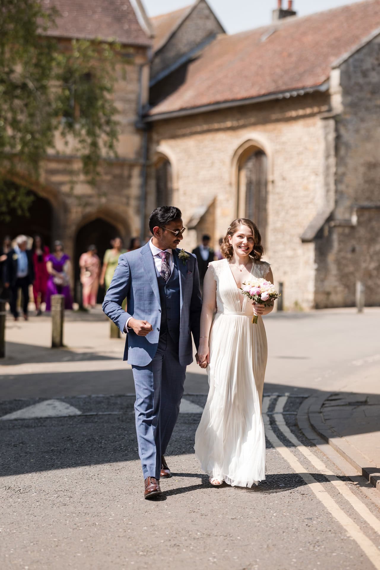A couple walks hand in hand, smiling at each other. The man wears a blue suit, and the woman wears a white dress, holding a bouquet. They are outdoors in a sunny area with historic stone buildings in the background.