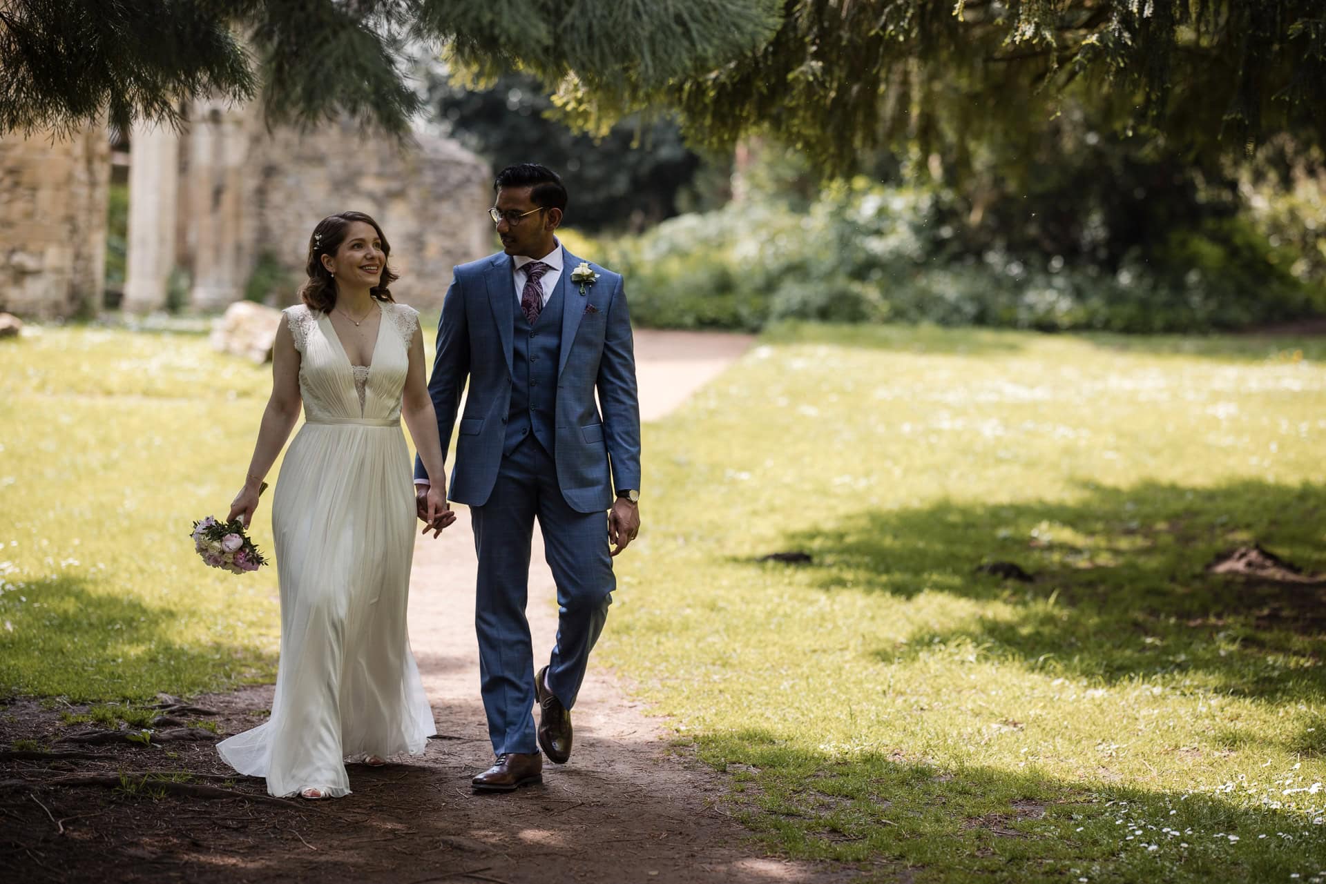 A couple in formal attire walk hand in hand along a sunlit garden path. The woman wears a white dress and holds a small bouquet, while the man wears a blue suit. Lush greenery surrounds them, creating a serene and romantic setting.