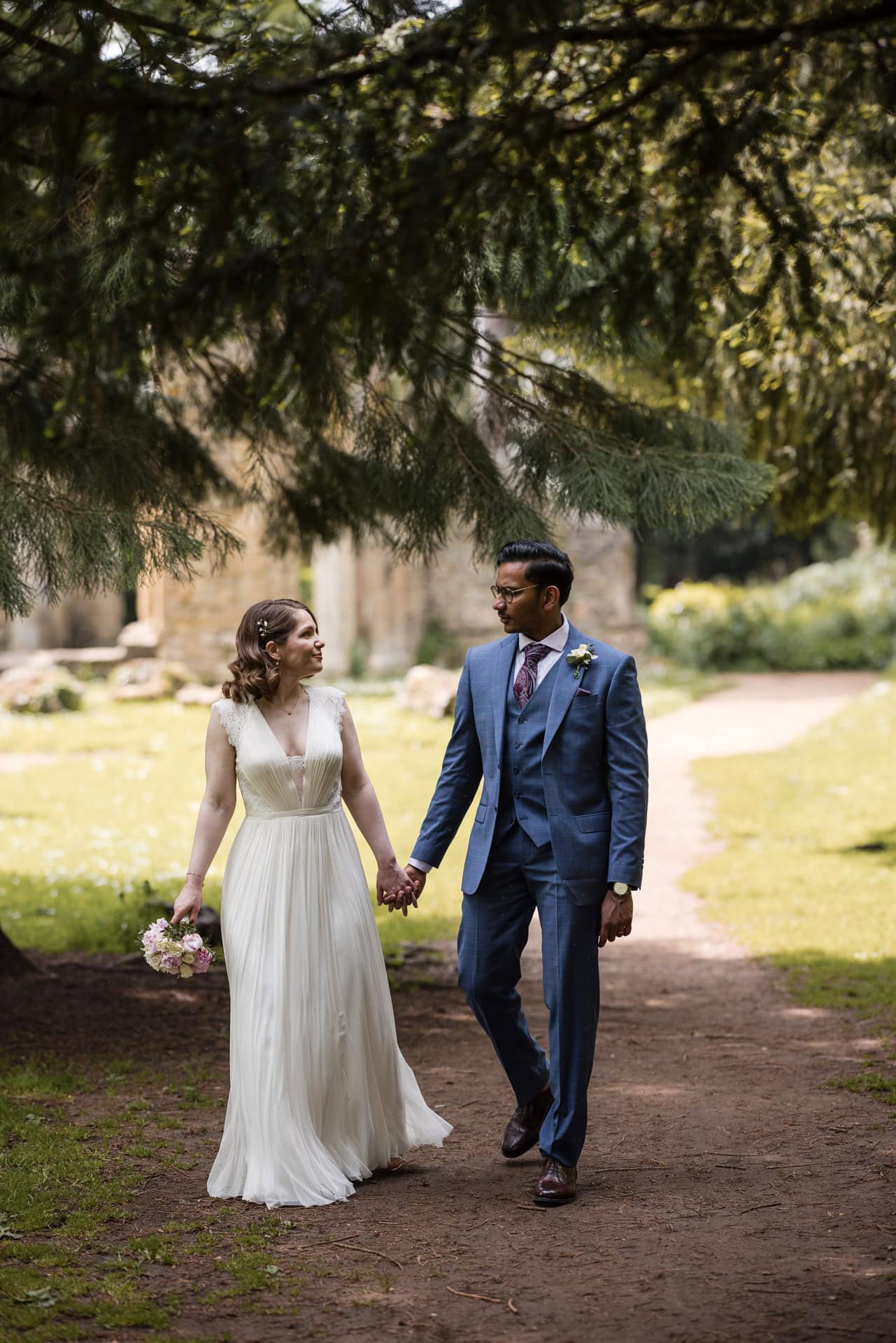 A bride in a white dress and a groom in a blue suit walk hand in hand on a shaded path under a tree. They are outdoors, with greenery and sunlight in the background, looking at each other and smiling.
Oxford Wedding Photographer