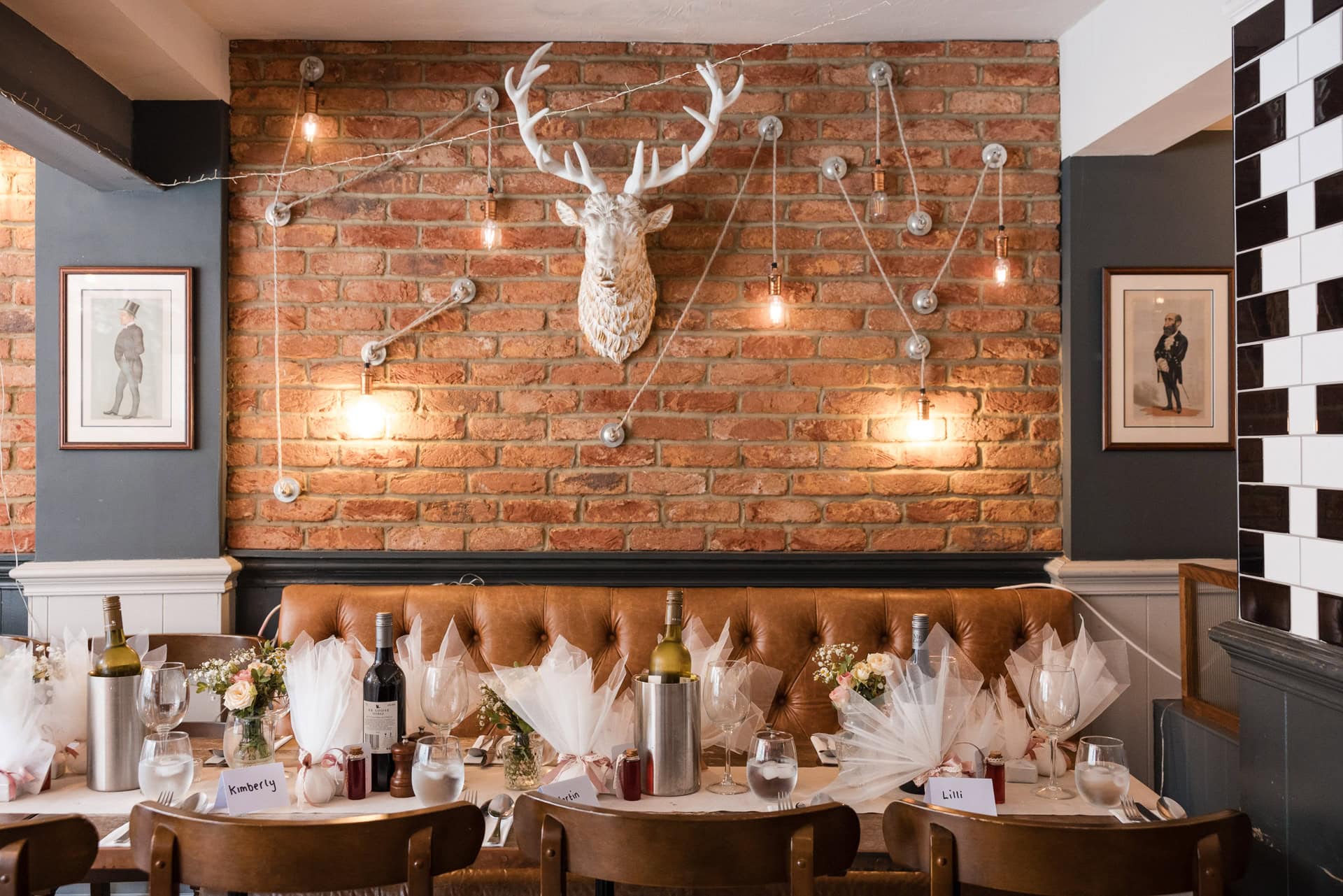 Cozy restaurant interior with a brick wall featuring a decorative deer head and string lights. Tables are set with wine bottles, glasses, and gift bags. Simple decorations create a warm atmosphere.