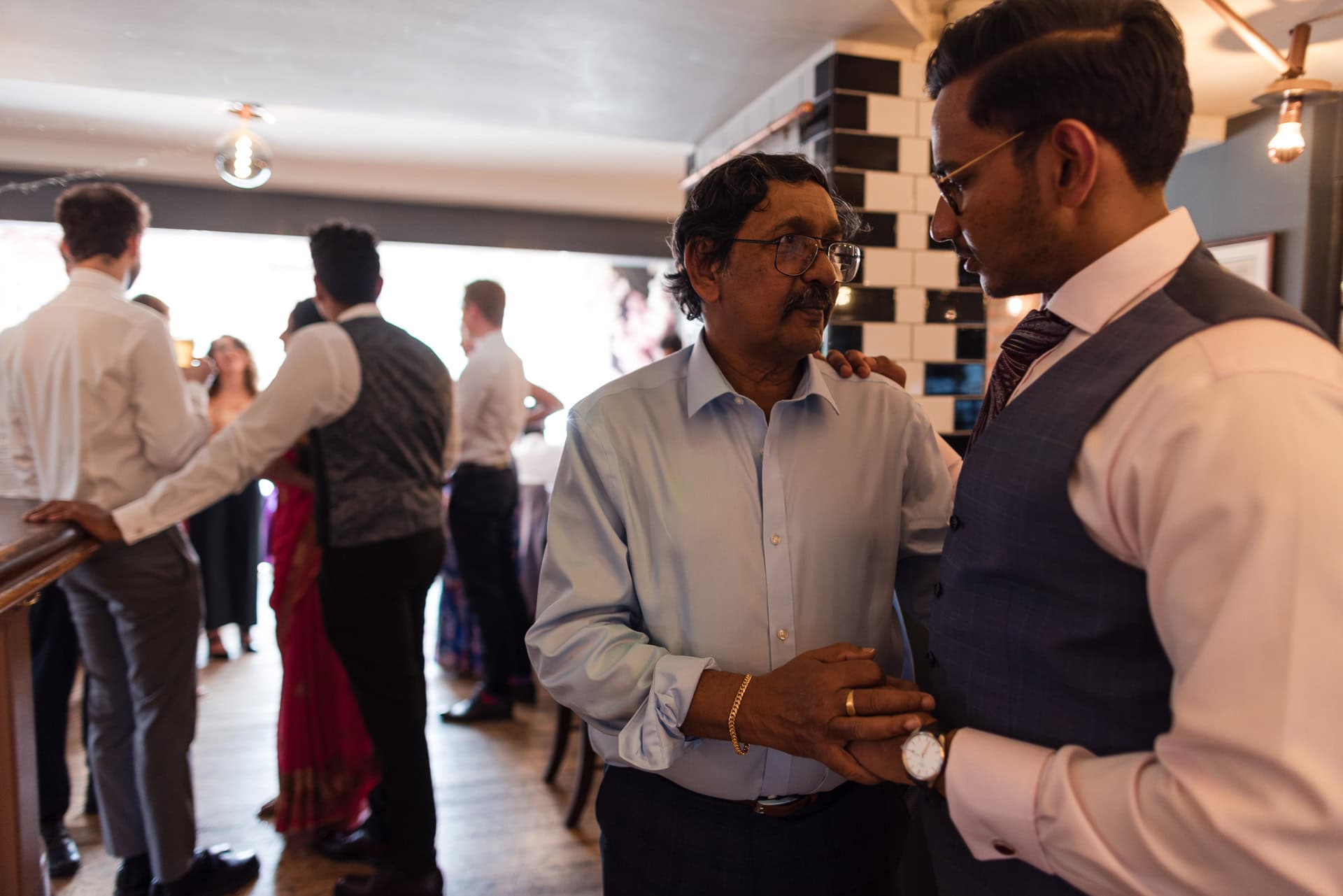 Two men in conversation at a social gathering, one in a light blue shirt and the other in a vest and tie. They stand near a tiled wall. Others are mingling in the background, dressed in formal attire. The setting appears to be indoors, possibly a reception.
