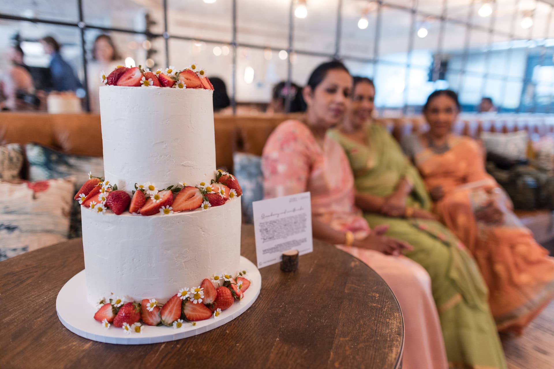 A two-tier white cake adorned with strawberries and small flowers sits on a wooden table. In the blurred background, three women wearing colorful dresses are seated on a couch.