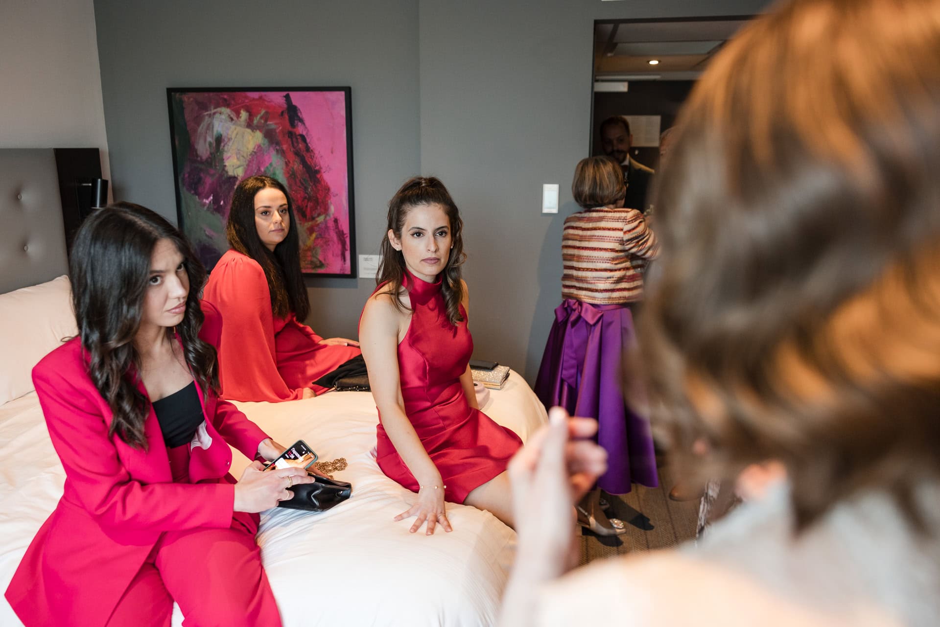A group of women in a room, dressed in red and pink formal attire, are sitting and standing around a bed. One is holding a mobile phone. A painting with pink hues is on the wall. Another person is blurred in the foreground.