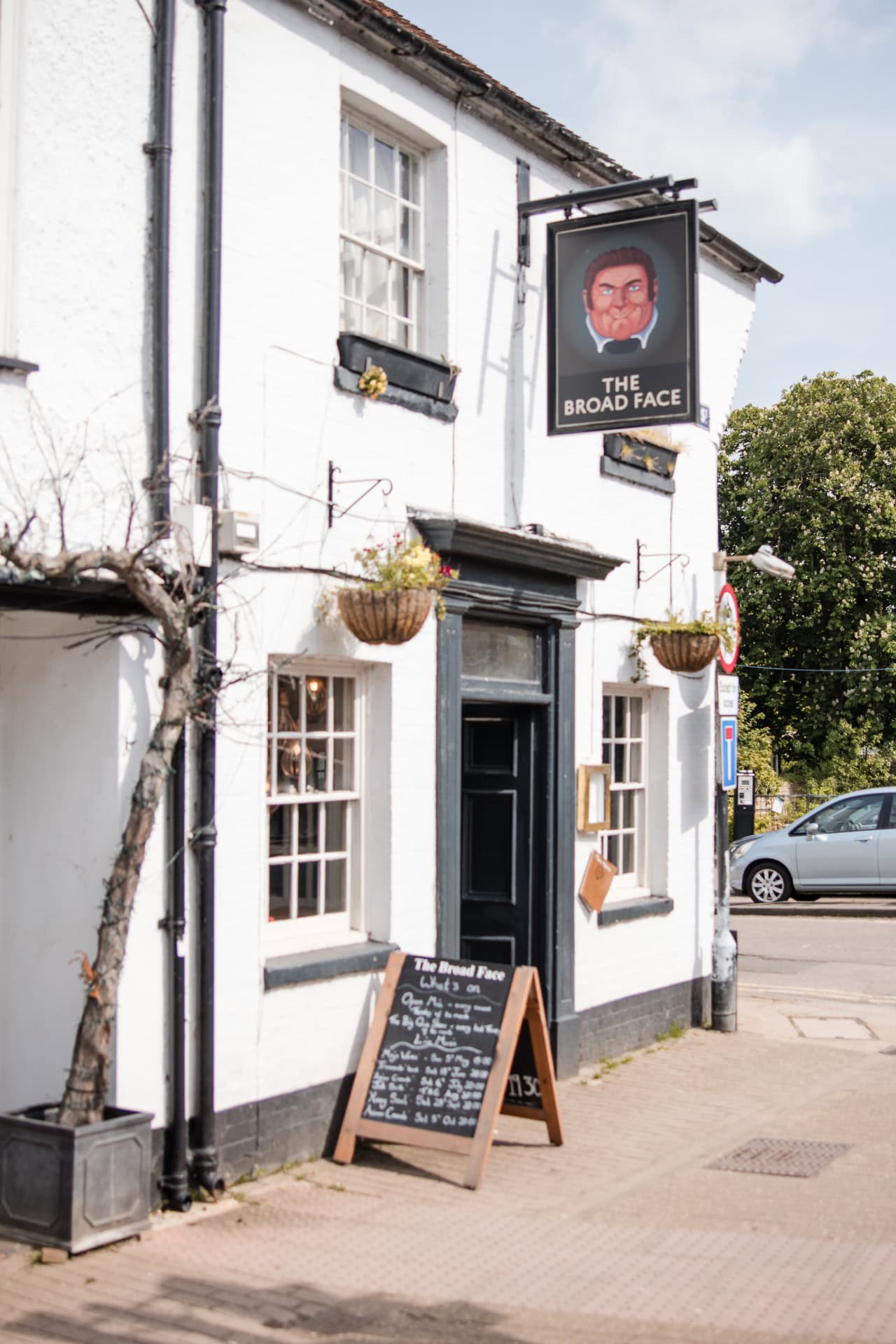 The image shows a white building with a dark door and windows, featuring a pub sign reading "The Broad Face" with an illustration. Hanging baskets adorn the facade, and a chalkboard menu leans against the wall. A car is parked nearby.