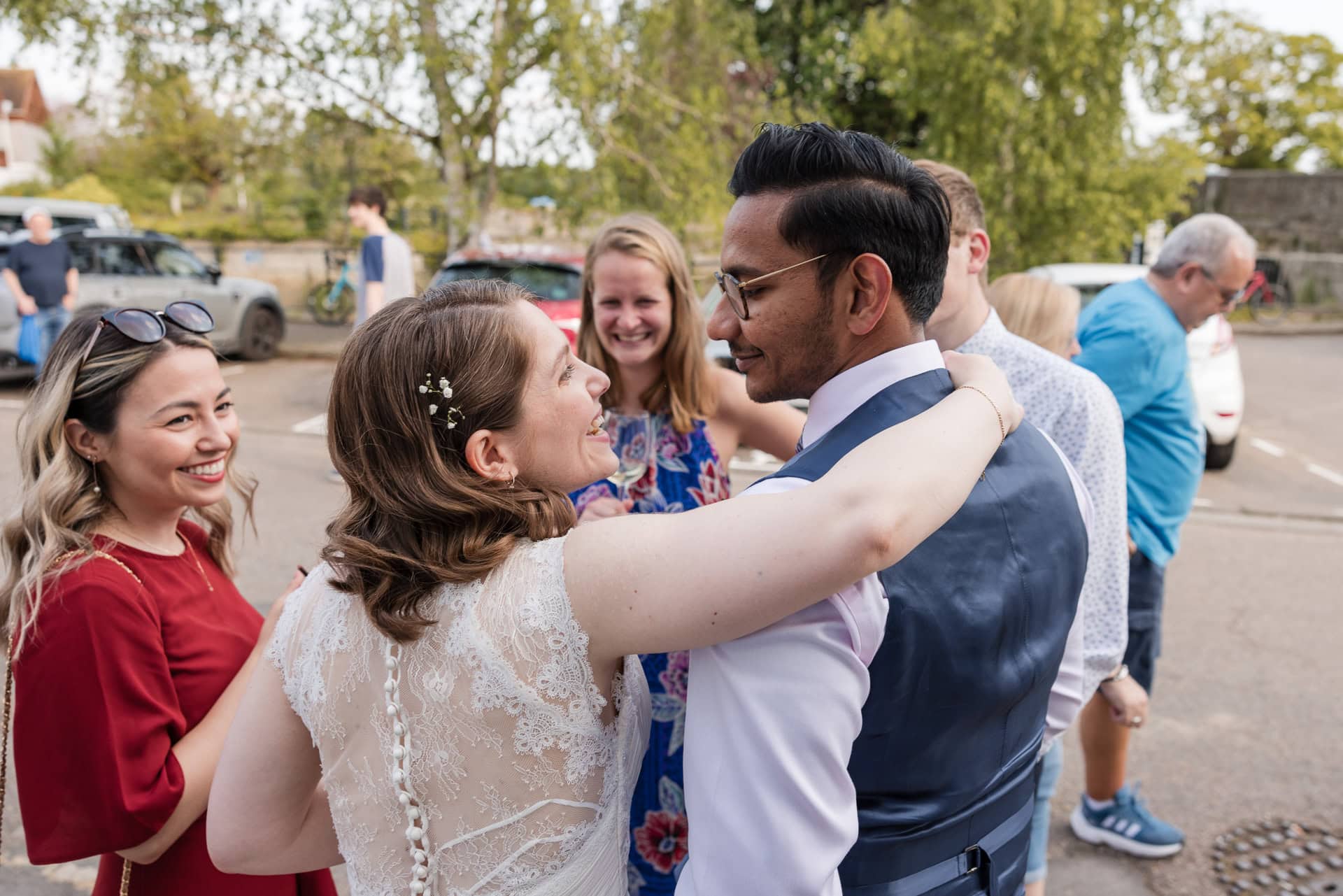 A group of people are outside in a parking lot, smiling and chatting. A woman in a white dress with floral decorations embraces a man in a vest and glasses. Others around them appear to be enjoying the moment together.