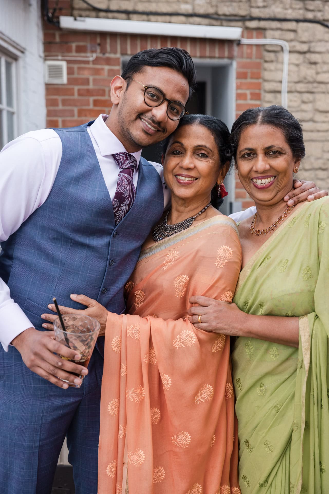 A man in a suit stands smiling with two women in traditional attire. One woman is in an orange sari and the other in a green sari. They are outdoors, near a brick wall, looking happy and posing together.