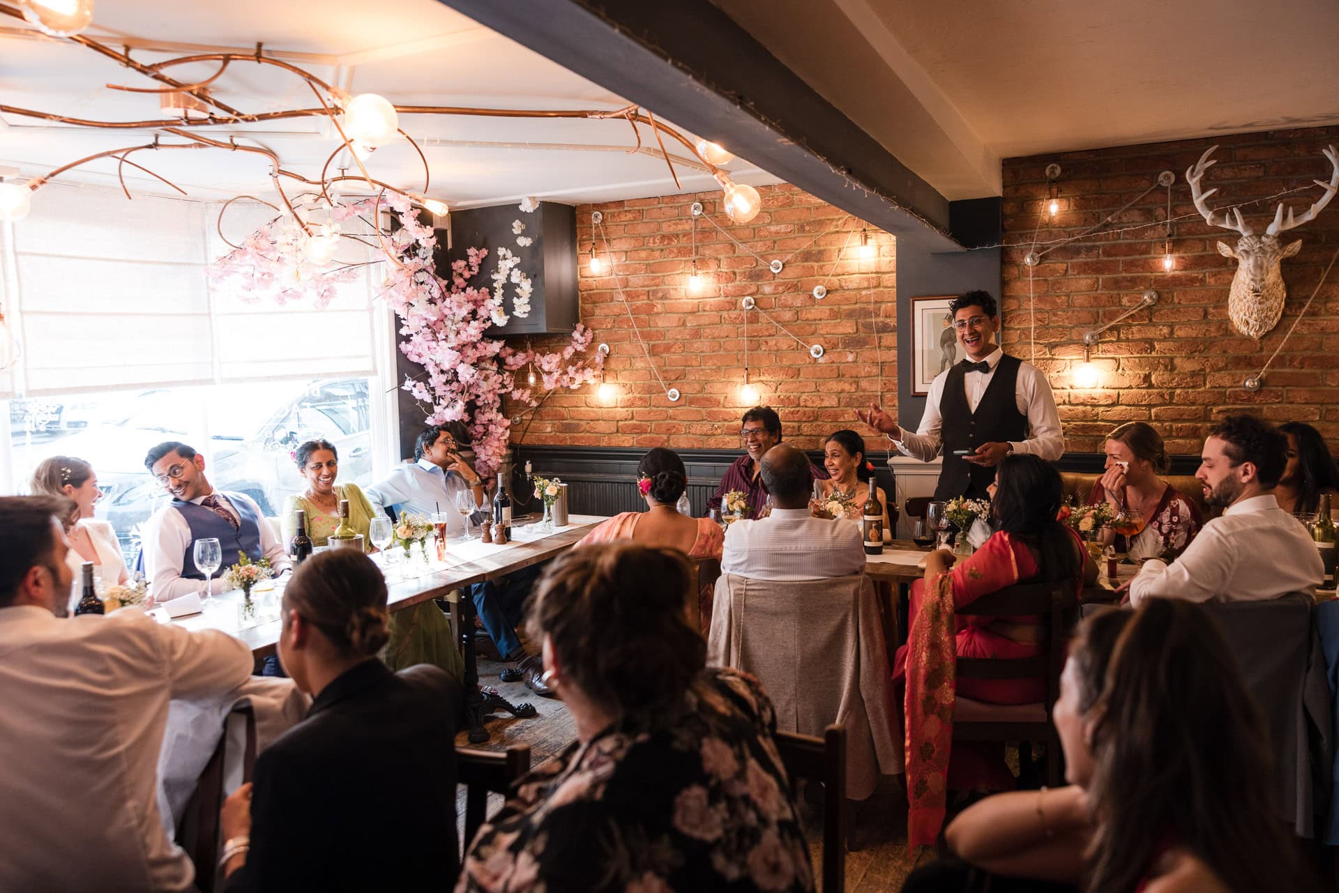 A group of people seated at a long table in a warmly lit restaurant with brick walls and decorative branches. A waiter stands at one end, speaking to the guests, who are engaged in conversation and laughter. Tables are adorned with flowers and candles.
