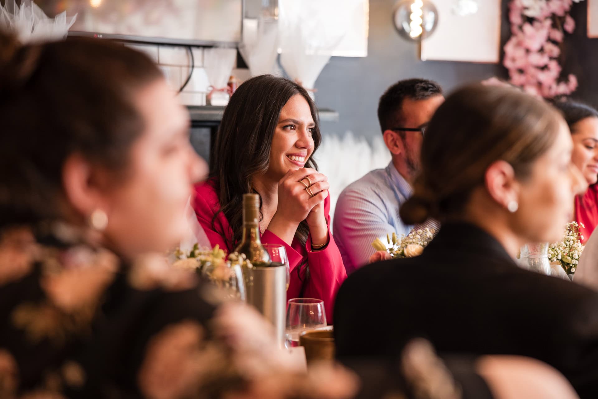 A group of people sitting together at a dining table, with a woman in a red top smiling and resting her chin on her hands. The table is decorated with flowers and a wine bottle. The background is softly blurred, creating a warm atmosphere.
