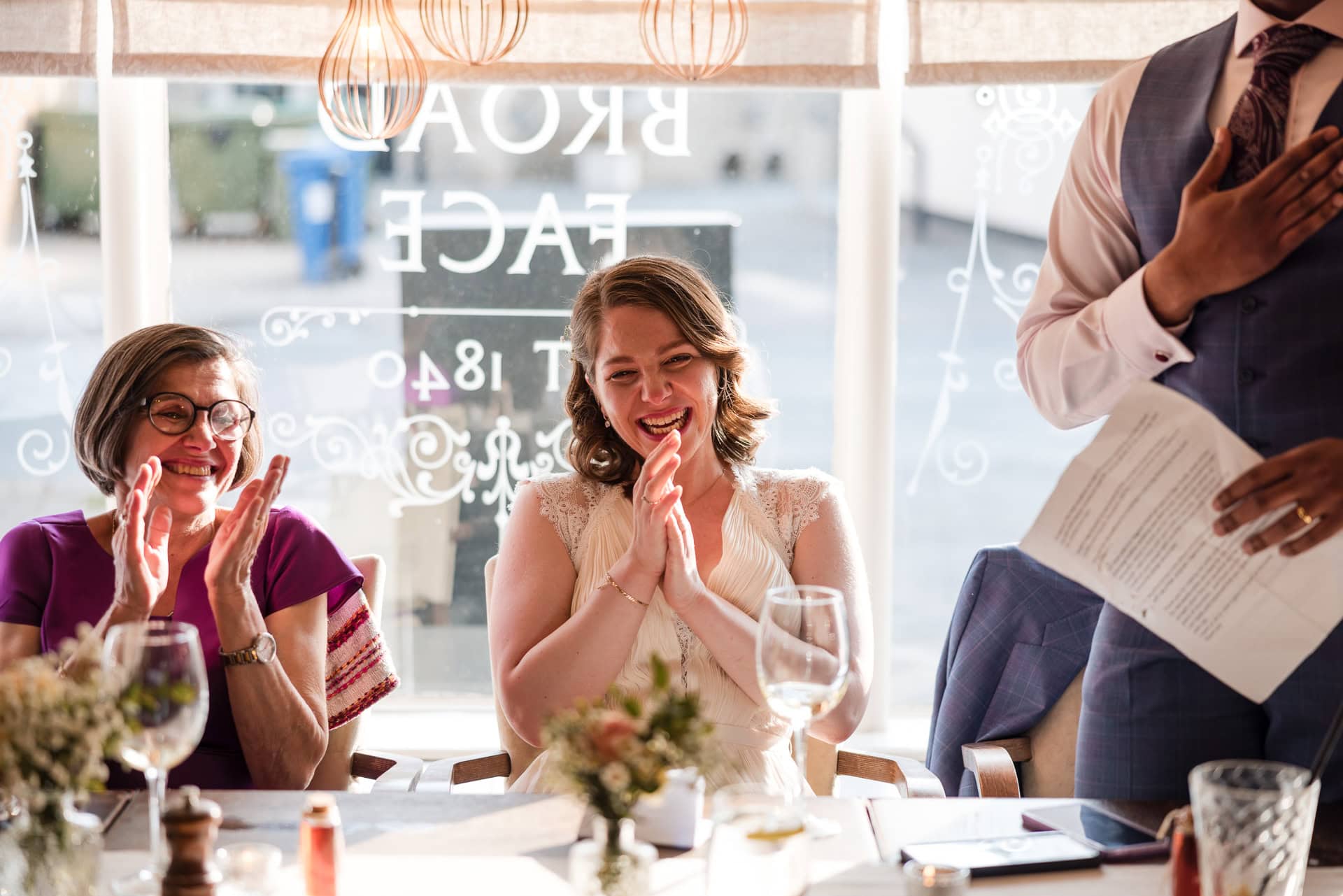 A group of people enjoying a celebration at a restaurant. Two women are seated, smiling and applauding. One is wearing glasses and a purple dress, the other a light dress. A standing person in a suit gestures with a hand on their chest while holding papers.