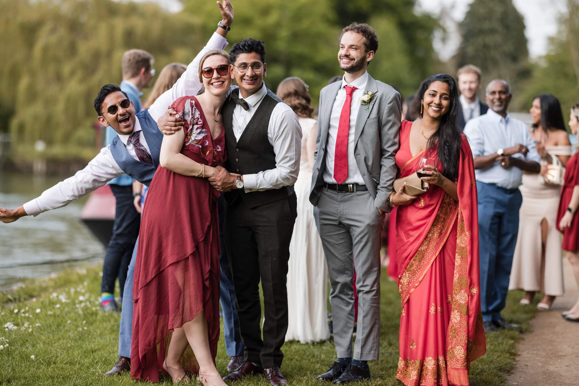 A group of people, some in formal and traditional attire, smiling outdoors by a river. A few are posing playfully, and the background is lush with green trees.