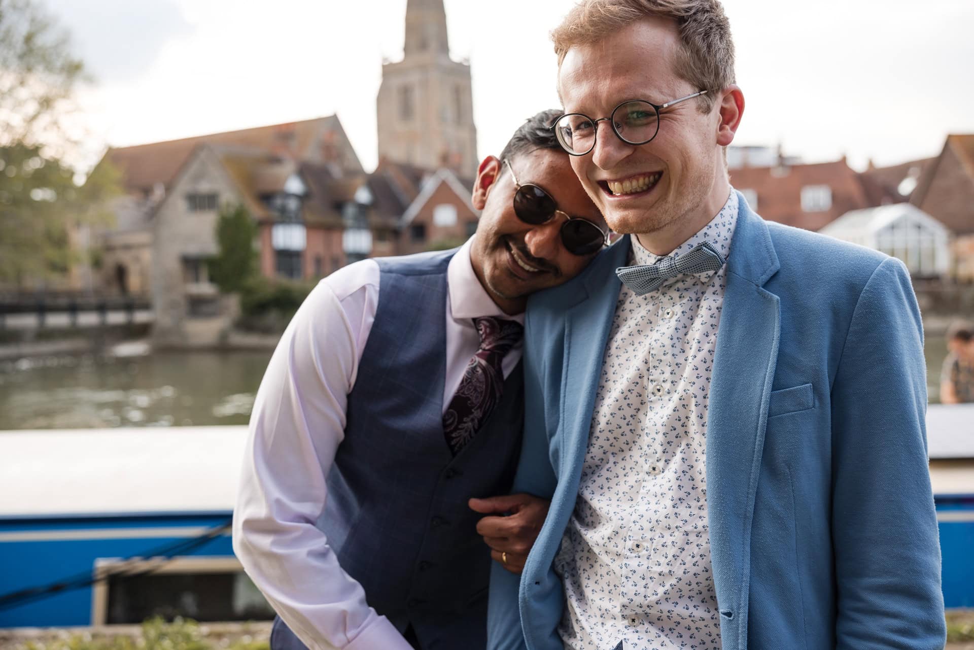 Two men laughing and smiling near a river. One man wears glasses and a blue jacket, while the other leans on his shoulder, wearing sunglasses and a vest. In the background are buildings and a church spire.