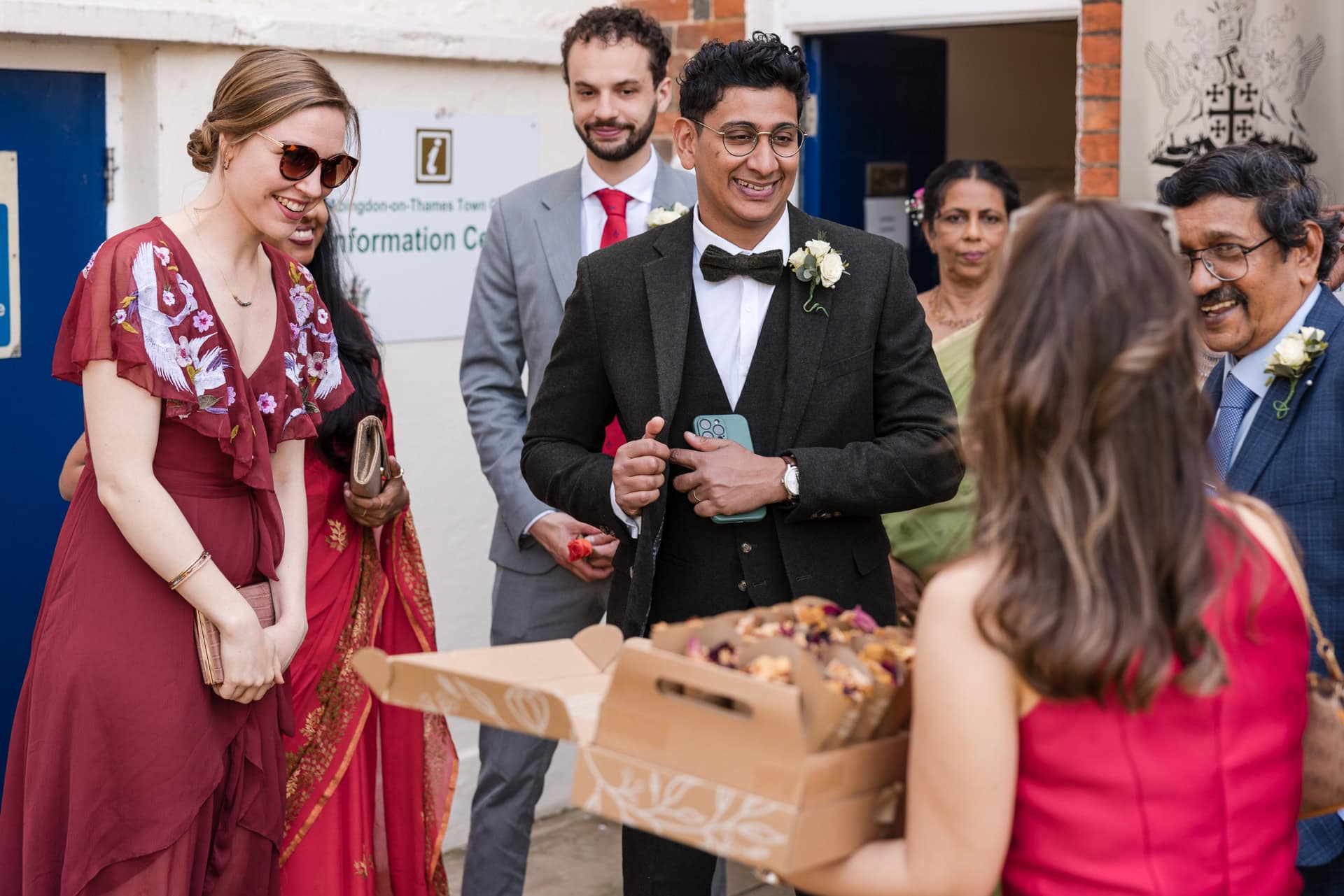 A group of people in formal attire stand together, smiling and interacting. One person holds a box of pastries. They are in front of a building entrance labeled "Information Centre." The atmosphere appears celebratory.