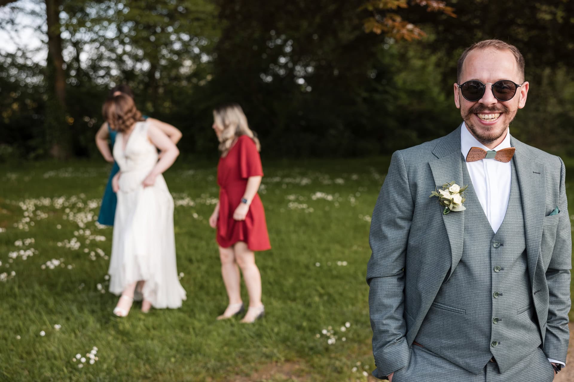 A man wearing sunglasses and a gray suit with a boutonniere smiles, standing on grass. In the background, three women in dresses are talking and adjusting clothing in a field with trees and daisies.