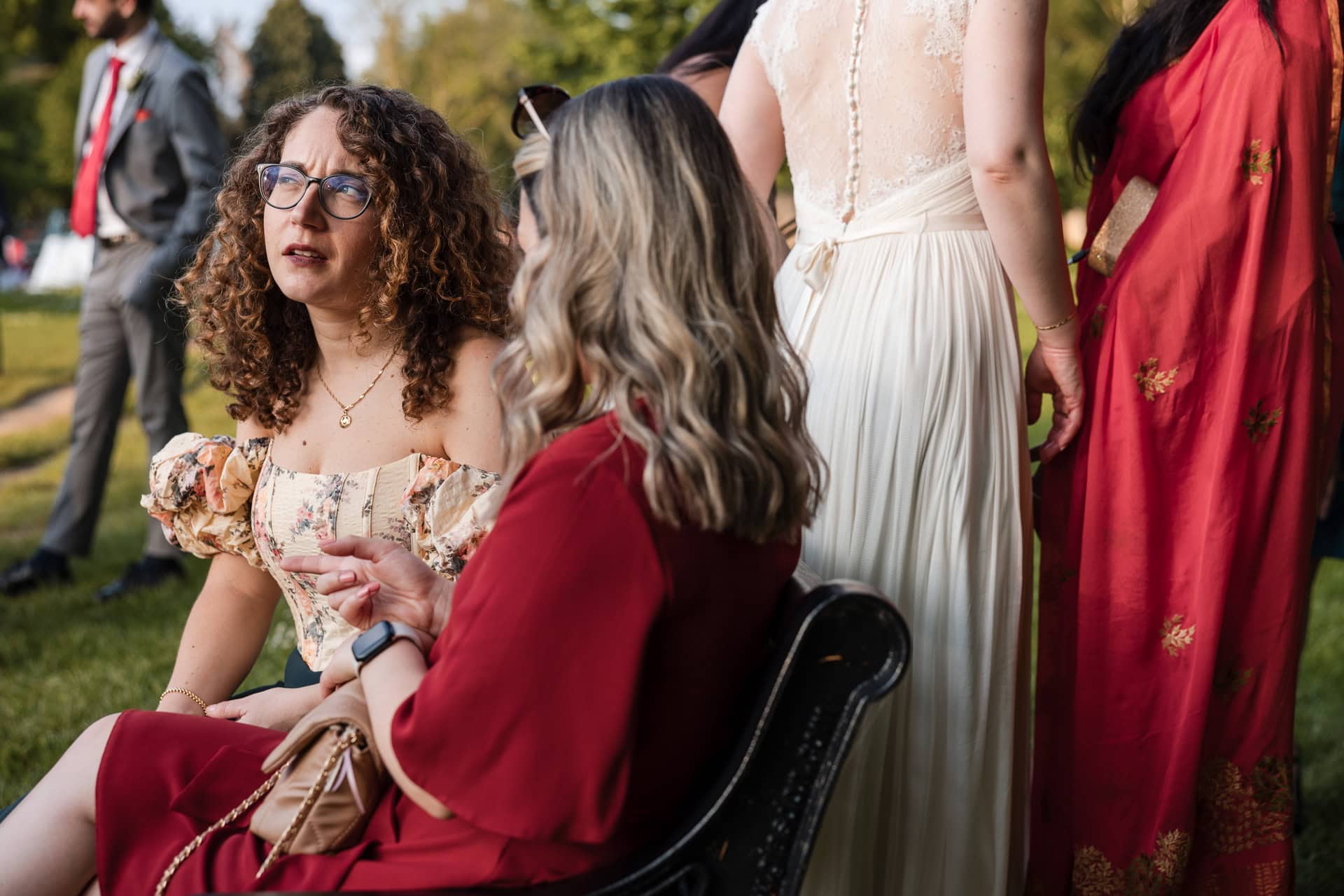 Two women sit on a bench outdoors, engaged in conversation. One wears glasses and a floral dress, the other a red outfit. In the background, people are dressed in formal attire, with trees and grass around them, suggesting a gathering.