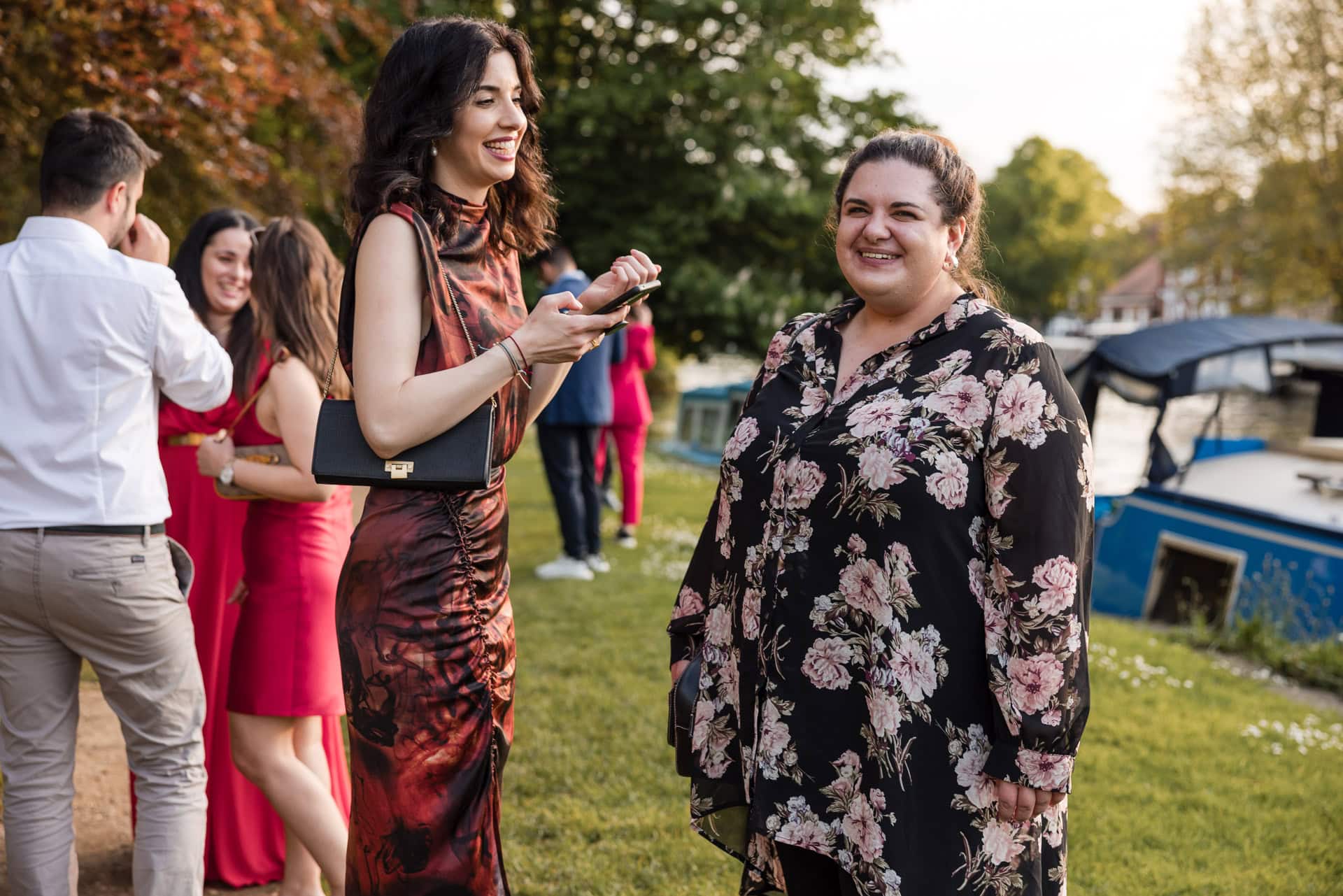Two women smiling and chatting near a riverbank, surrounded by others. One wears a floral black dress, and the other a patterned red dress. A boat is docked nearby, with trees and grass in the background.