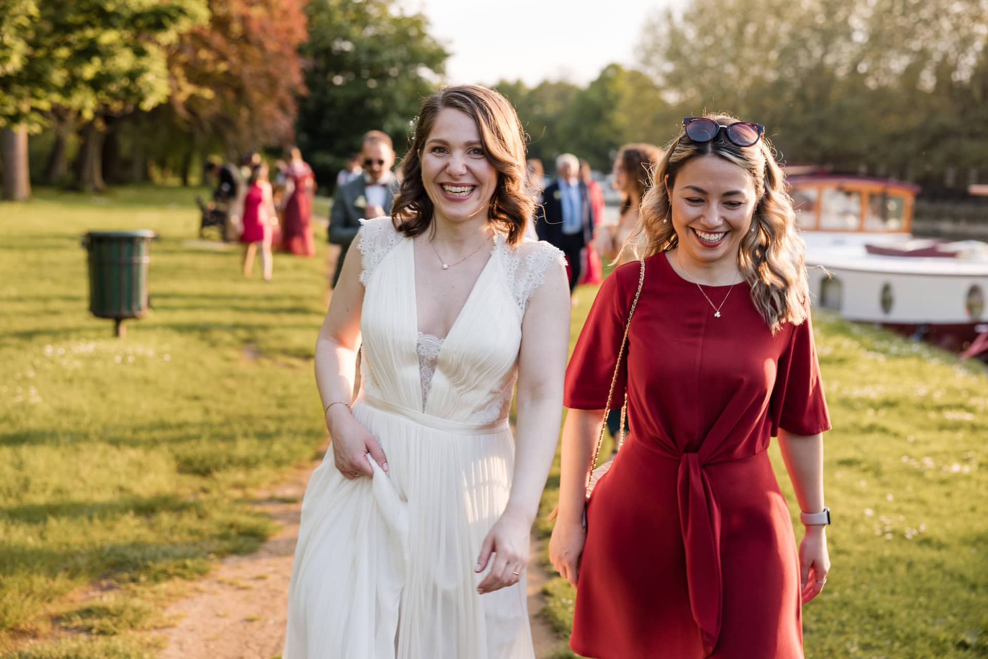 Two women walking on a grassy path, one in a white dress and one in a red dress, smiling and enjoying a sunny day. Trees and people are in the background. Oxford Wedding Photographer