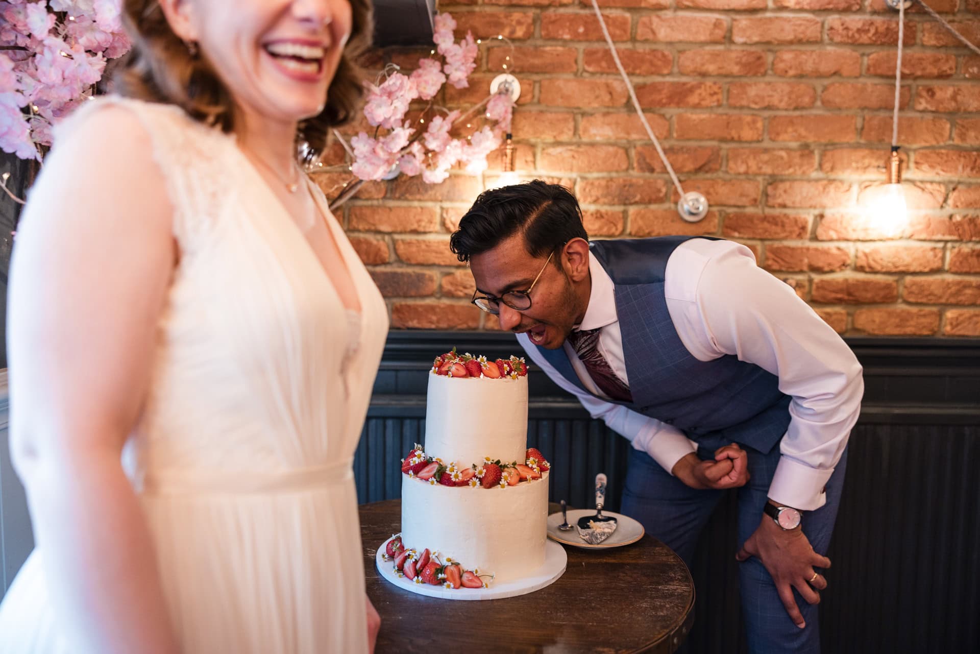 A man playfully pretends to take a bite out of a two-tiered cake decorated with strawberries, while a woman in a white dress laughs beside him. They are in a cozy room with brick walls and string lights.