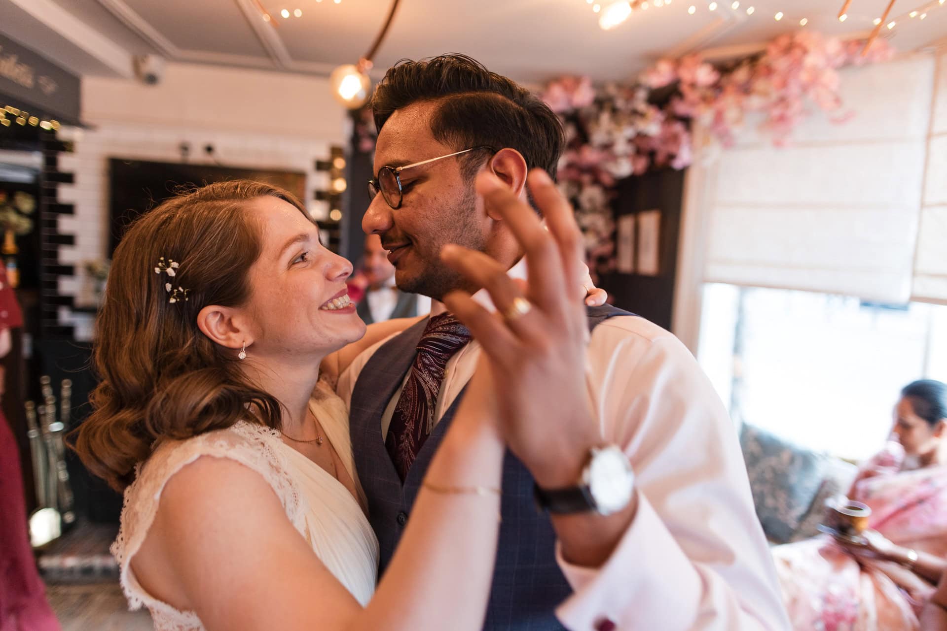 A happy couple dances closely, smiling at each other in a warmly lit room decorated with pink flowers. Other people are seated in the background, and some string lights add to the cozy atmosphere.