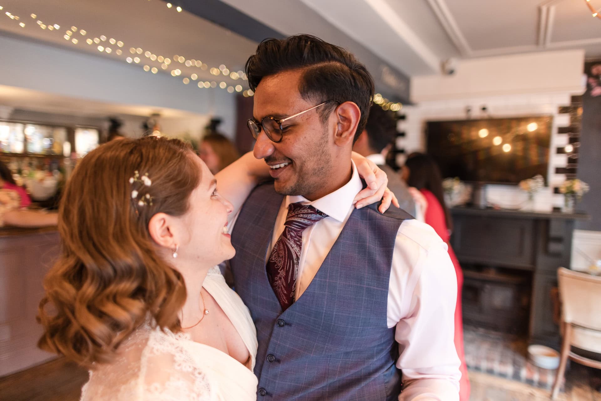 A couple dances closely, smiling at each other in a warmly lit room decorated with string lights. The woman wears a white dress with floral hair accessories, and the man wears glasses and a suit with a patterned tie.
Oxford Wedding Photographer