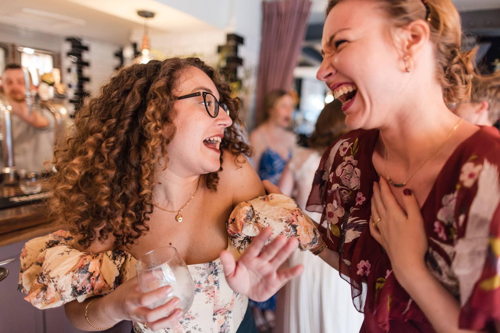 Two women are joyfully laughing in a social setting. One is holding a drink, wearing a floral dress and glasses, while the other, also in a floral outfit, has her hand on her chest. Both are sharing a candid, happy moment indoors.Oxford Wedding Photographer