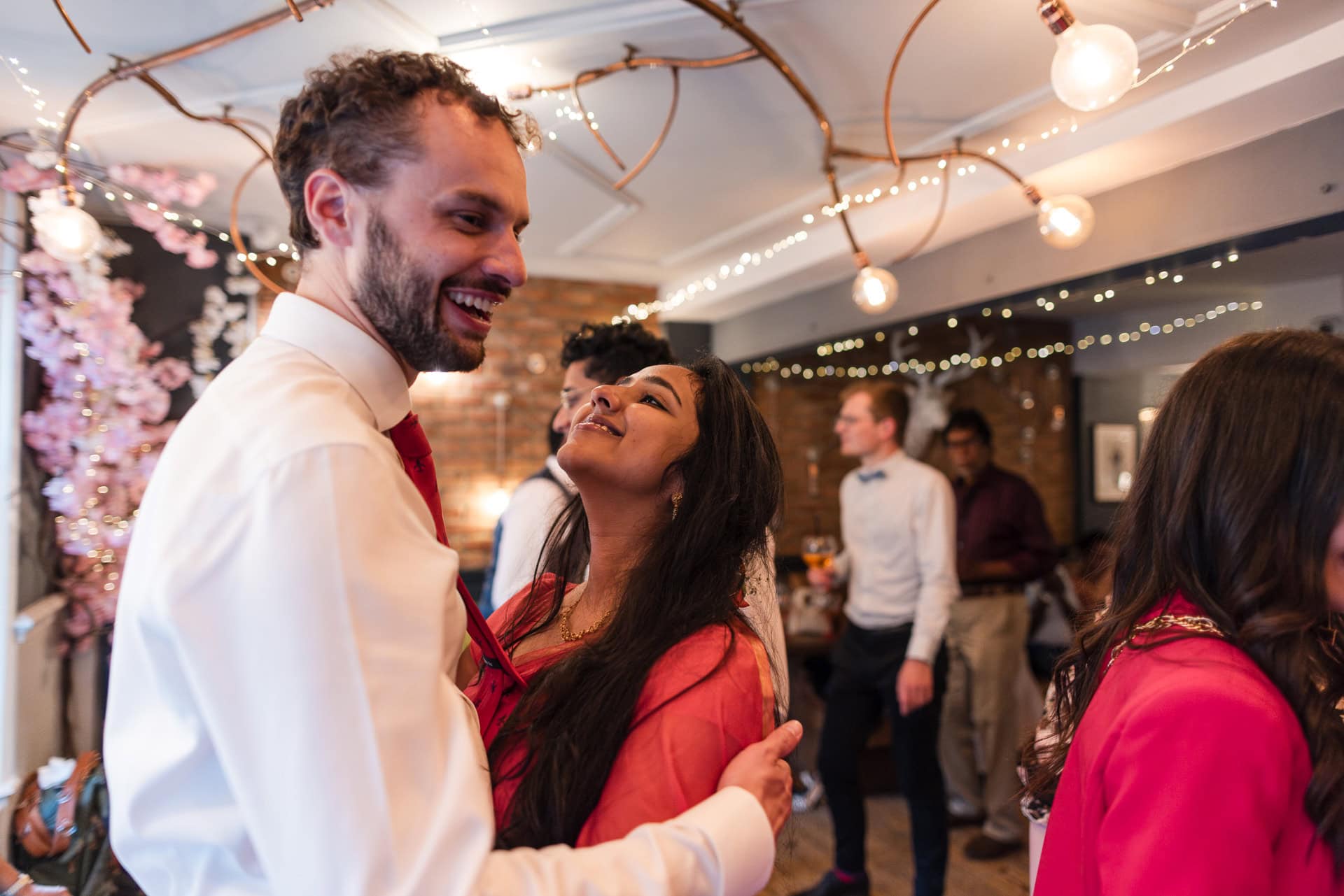A man and woman smile and embrace in a warmly lit room with decorative string lights and flowers. Other guests converse and mingle in the background, dressed in formal attire.