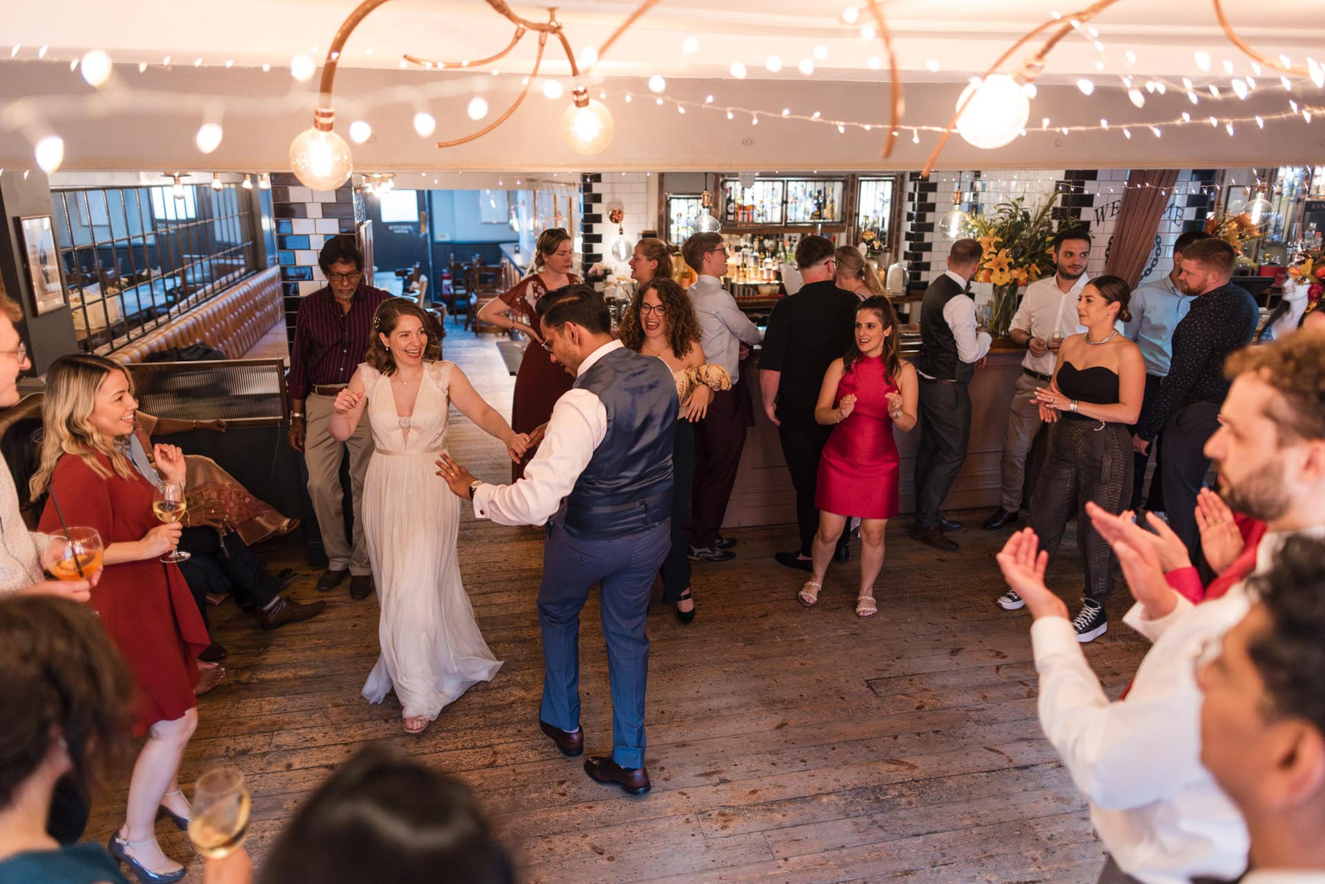 A wedding reception in a warmly lit venue with string lights. A bride and groom dance joyfully in the center, surrounded by applauding guests dressed in formal attire. The scene is lively and celebratory, with people smiling and chatting. Oxford Wedding Photographer